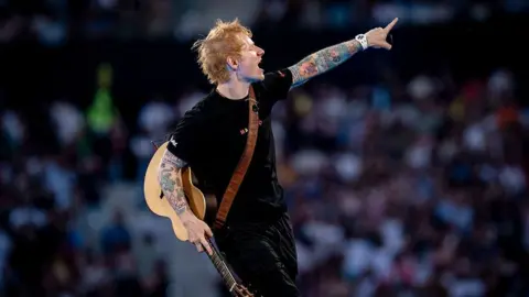 A. Perez Meca/ Getty Images Ed Sheeran, holding his guitar, points out at the crowd, which can be seen blurry in the background.